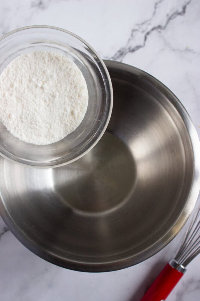 A glass bowl with white flour is held above an empty metal mixing bowl on a marble countertop; a whisk with a red handle lies nearby.