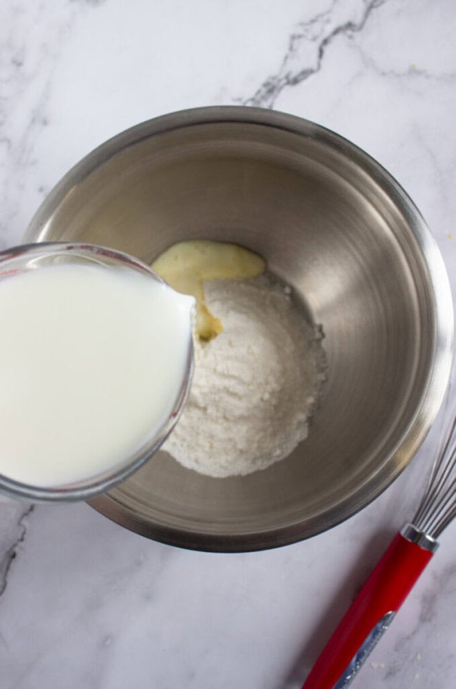 A measuring cup pours milk into a mixing bowl containing flour and butter, with a whisk placed nearby on a marble countertop.