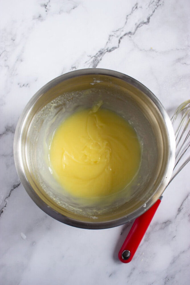 A stainless steel mixing bowl with a yellow batter inside, next to a metal whisk with a red handle on a white marble surface.