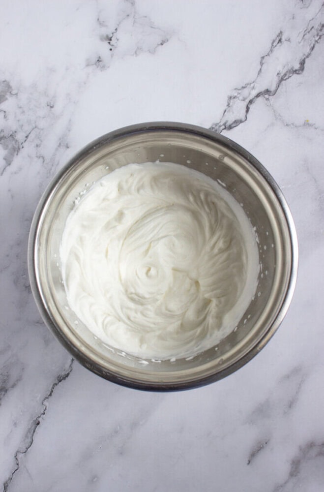 A stainless steel bowl filled with whipped cream sits on a white marble surface.