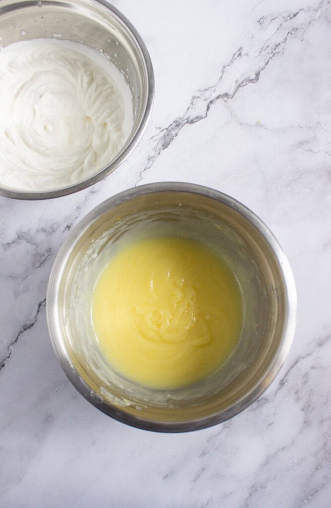 Two metal bowls on a marble surface; one contains whipped cream, and the other contains a yellow custard mixture.