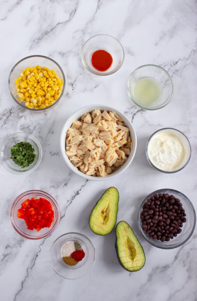 Nine bowls with ingredients for a recipe, including shredded chicken, corn, black beans, avocado, chopped red pepper, cilantro, spices, hot sauce, lime juice, and yogurt, on a marble surface.
