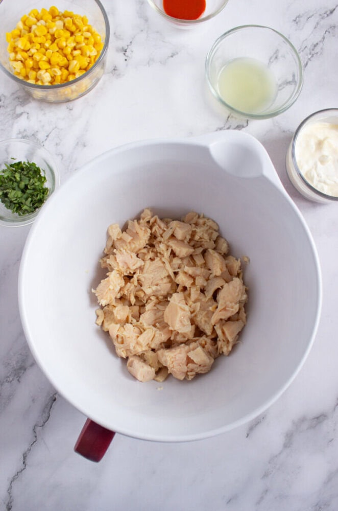 A white mixing bowl with canned tuna chunks, surrounded by bowls of corn, chopped herbs, mayonnaise, lime juice, and hot sauce on a marble surface.