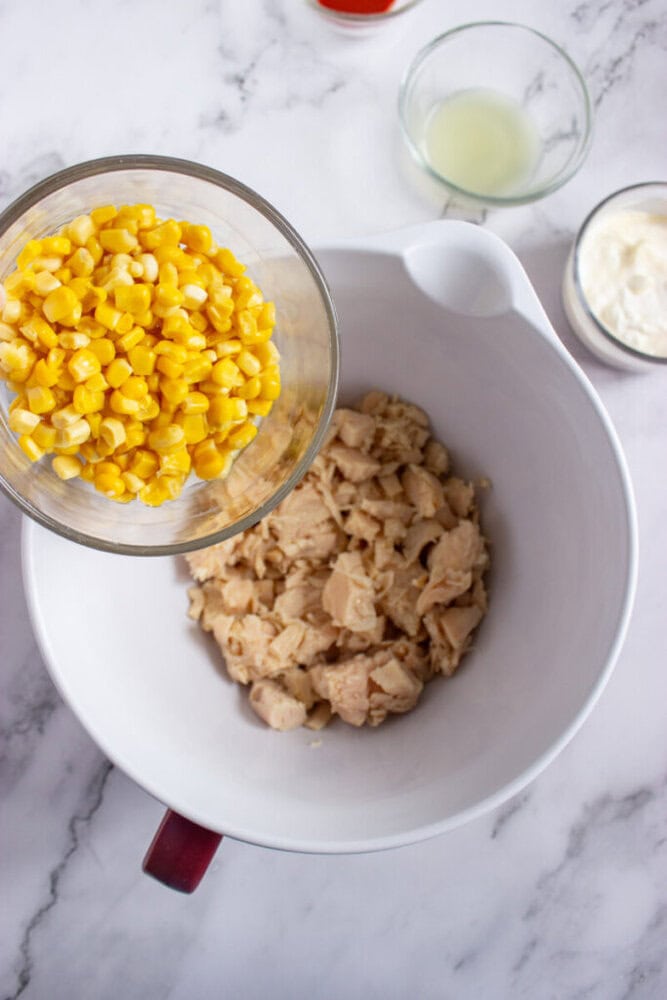 A glass bowl of corn is being poured into a large mixing bowl containing cooked, chopped chicken. Two small bowls with liquid and a creamy ingredient are nearby on a marble countertop.