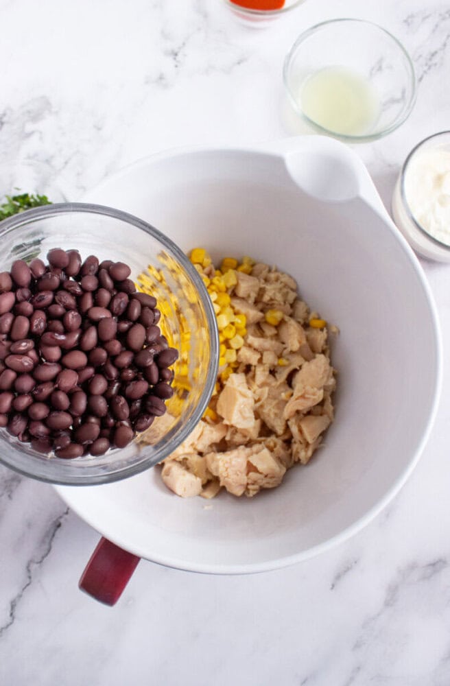 A glass bowl of black beans is being added to a mixing bowl containing cooked chicken and corn on a marble countertop.