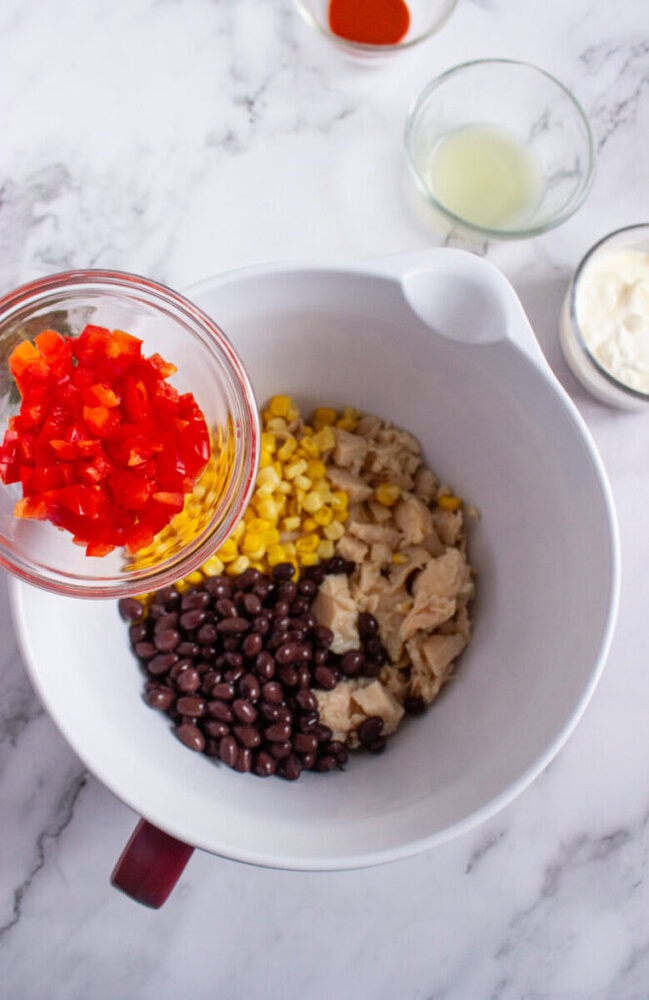 A bowl with black beans, corn, and chicken, with chopped red bell pepper being added; small bowls of sauce, lime juice, and mayonnaise are nearby.