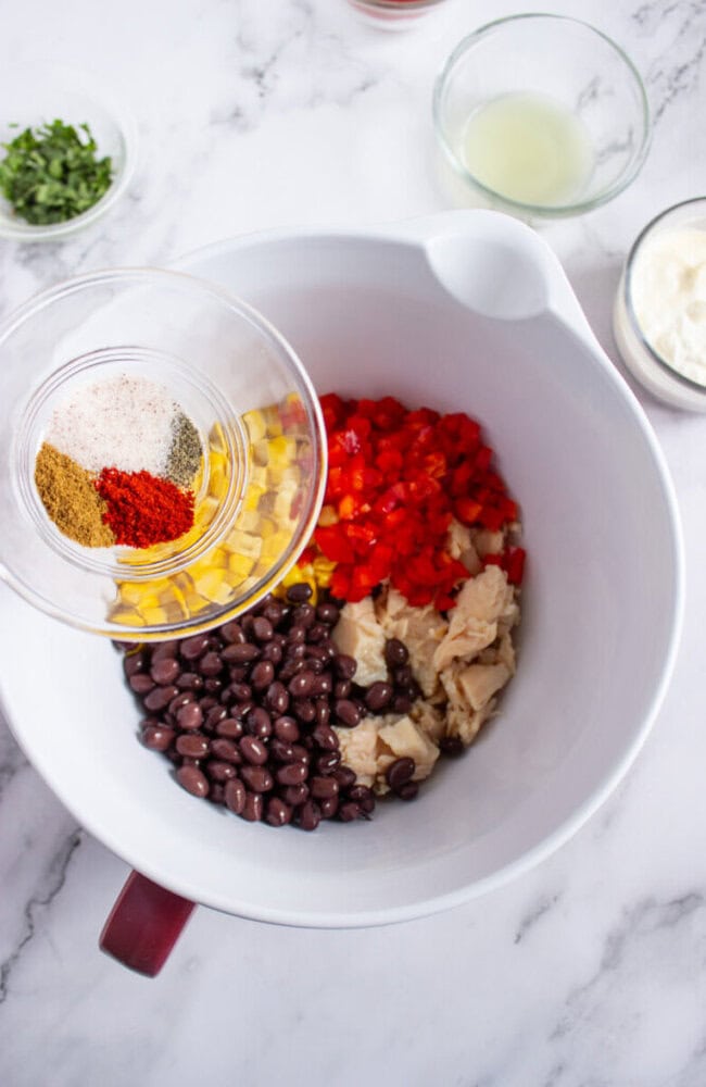 A mixing bowl contains black beans, corn, chopped red peppers, and chicken with a bowl of spices being added; other ingredients are in small bowls nearby on a marble countertop.
