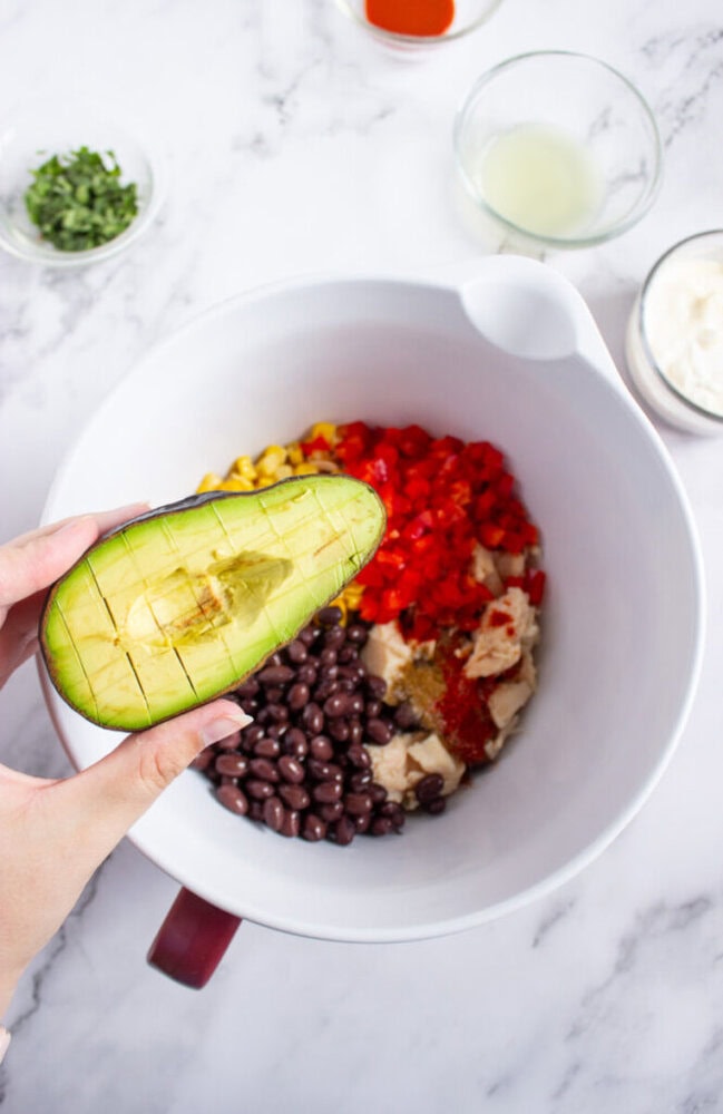 A hand holds a halved avocado over a mixing bowl containing black beans, corn, chopped red peppers, chicken, and spices. Small bowls of chopped herbs, lime juice, and sauce are nearby.