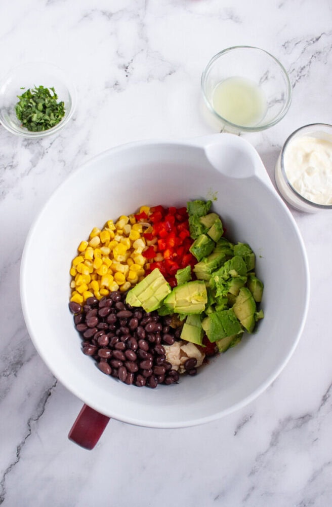 A mixing bowl with black beans, corn, diced red bell pepper, avocado, and seasonings. Nearby are bowls with chopped herbs, lime juice, and a white creamy sauce.