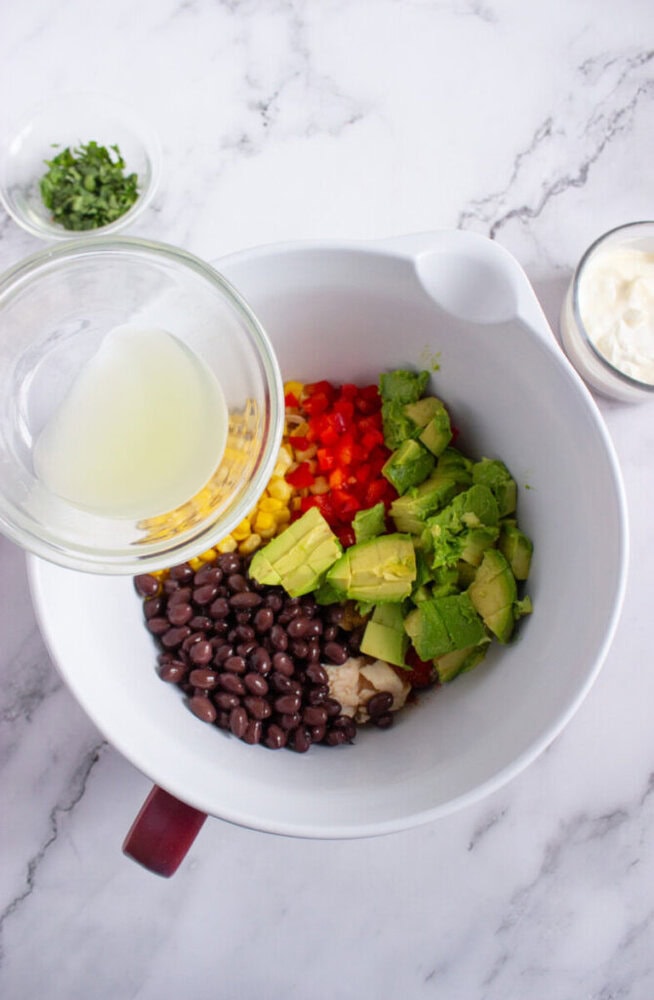 A mixing bowl with black beans, avocado, corn, red bell pepper, and herbs; a small bowl pours liquid in, with cilantro and a jar of sauce nearby on a marble surface.