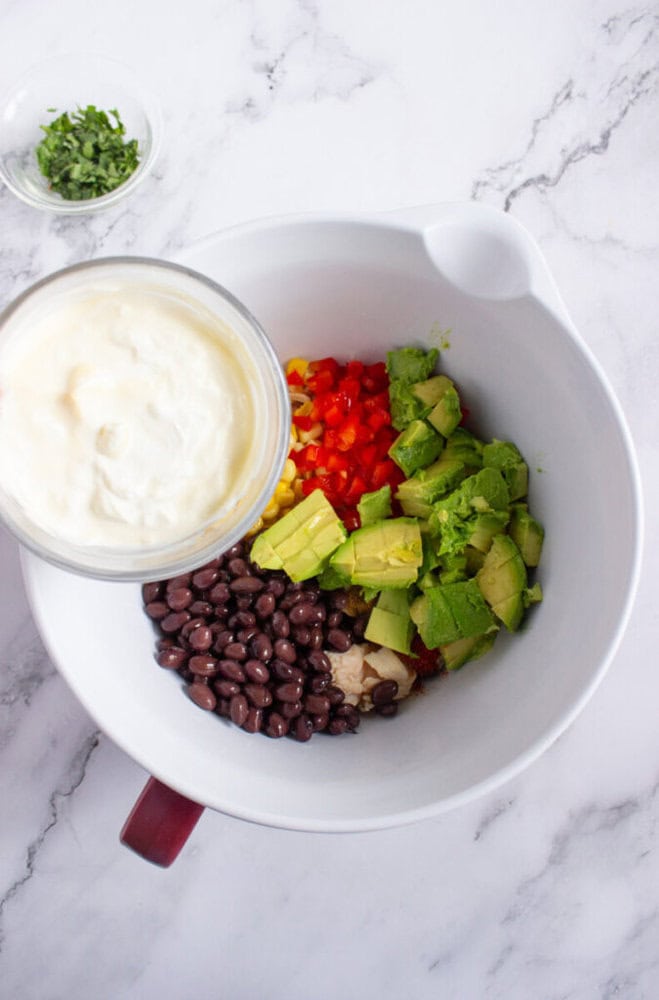 A white mixing bowl with black beans, chopped avocado, red bell pepper, corn, and diced onions. A separate bowl of mayonnaise and a small dish of chopped herbs are nearby.