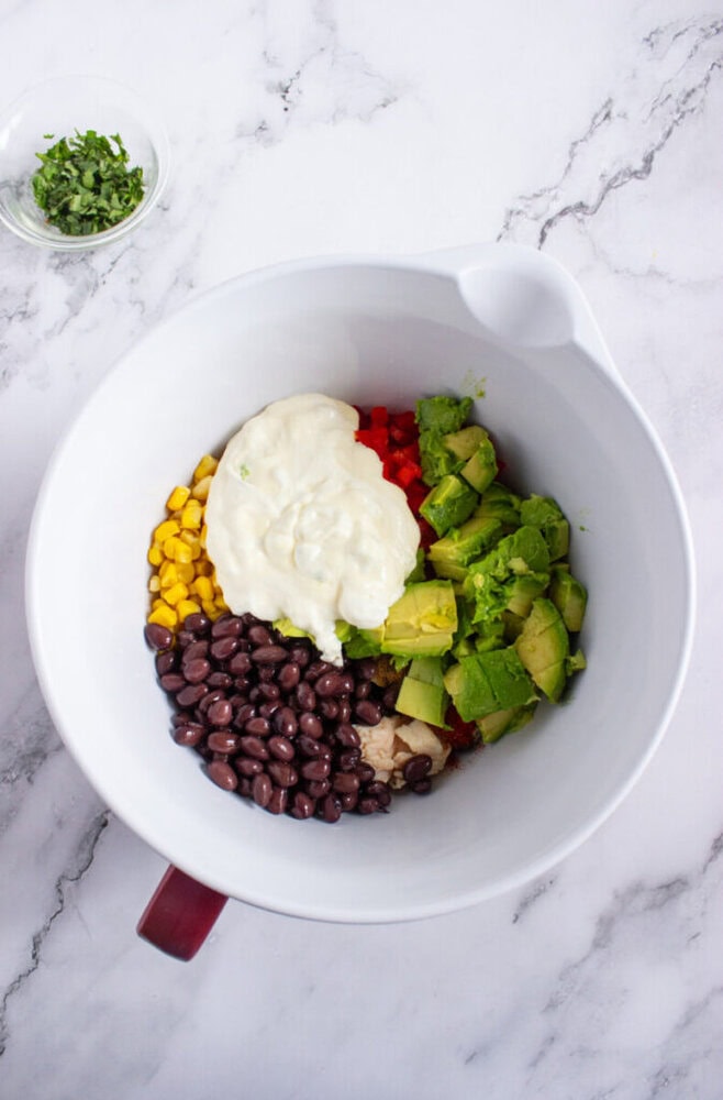 A large white bowl contains black beans, corn, chopped avocado, diced red pepper, and a dollop of creamy dressing. A small dish of chopped herbs sits nearby on a marble surface.