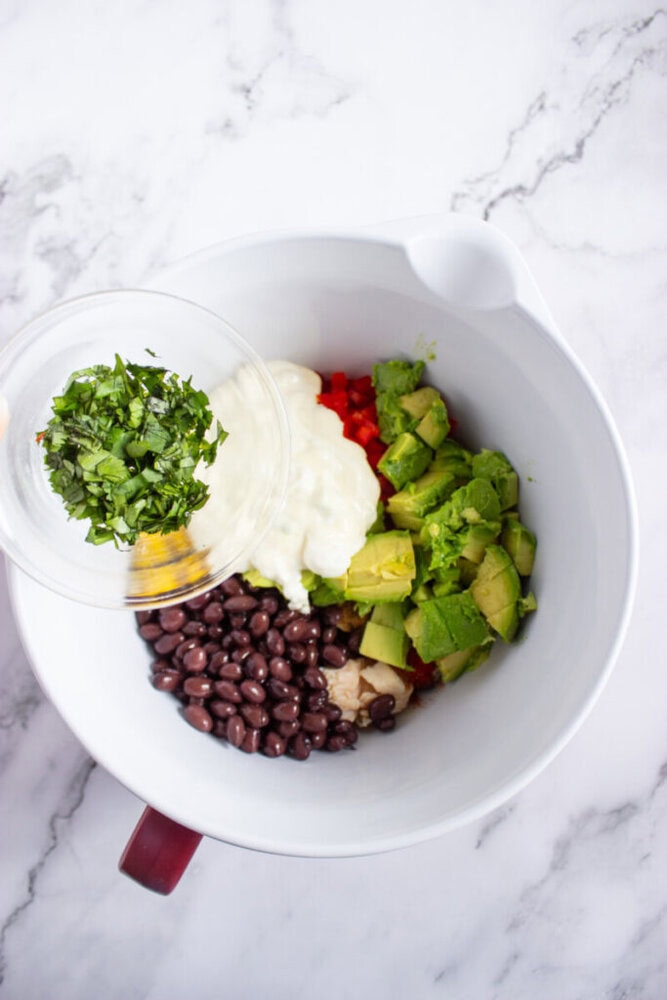 A mixing bowl containing black beans, diced avocado, red bell pepper, yogurt, and seasonings, with chopped cilantro being added from a small bowl above.