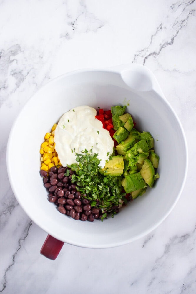 A white mixing bowl with black beans, corn, diced red bell peppers, avocado chunks, chopped cilantro, and a dollop of sour cream on a marble surface.