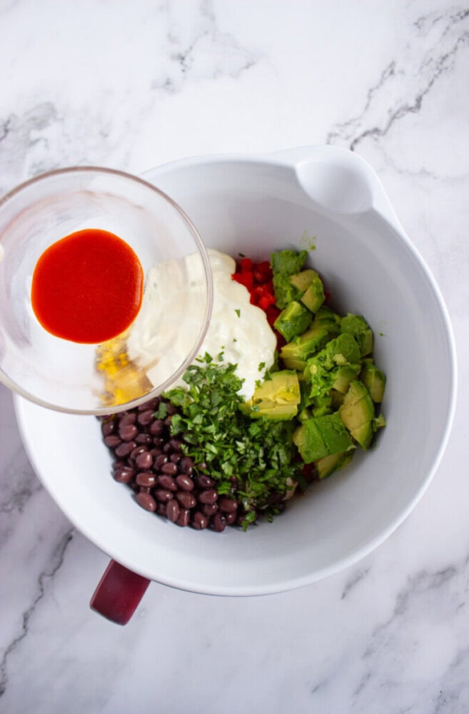 A mixing bowl with avocado, black beans, chopped herbs, diced red peppers, and mayonnaise, with a glass containing red sauce being poured in.