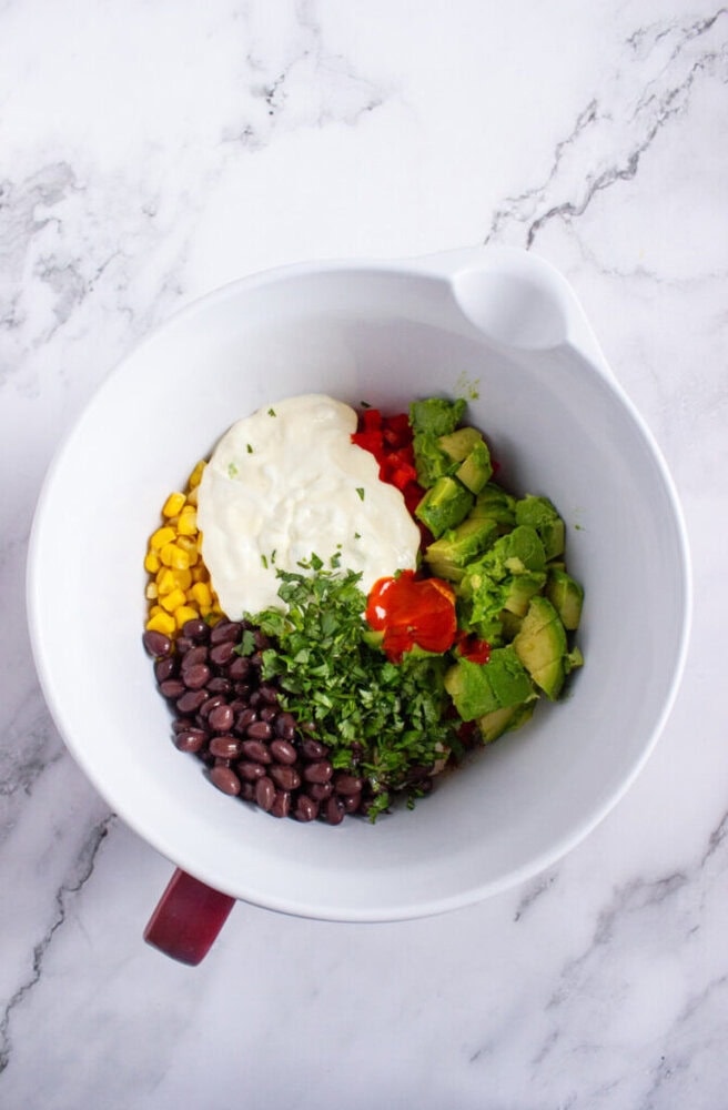 A white mixing bowl containing black beans, corn, diced avocado, chopped cilantro, diced red pepper, mayonnaise, and hot sauce on a marble surface.
