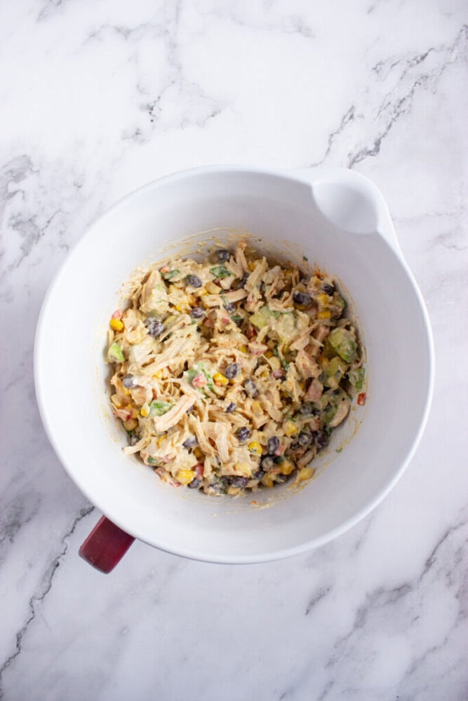 A white mixing bowl filled with a mixture of shredded chicken, black beans, corn, diced avocado, and other ingredients on a marble countertop.