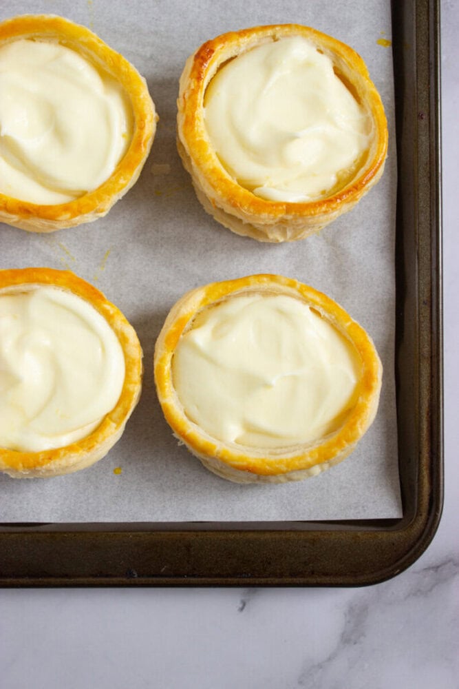 Four round pastry shells filled with creamy custard are arranged on a parchment-lined baking sheet, viewed from above.