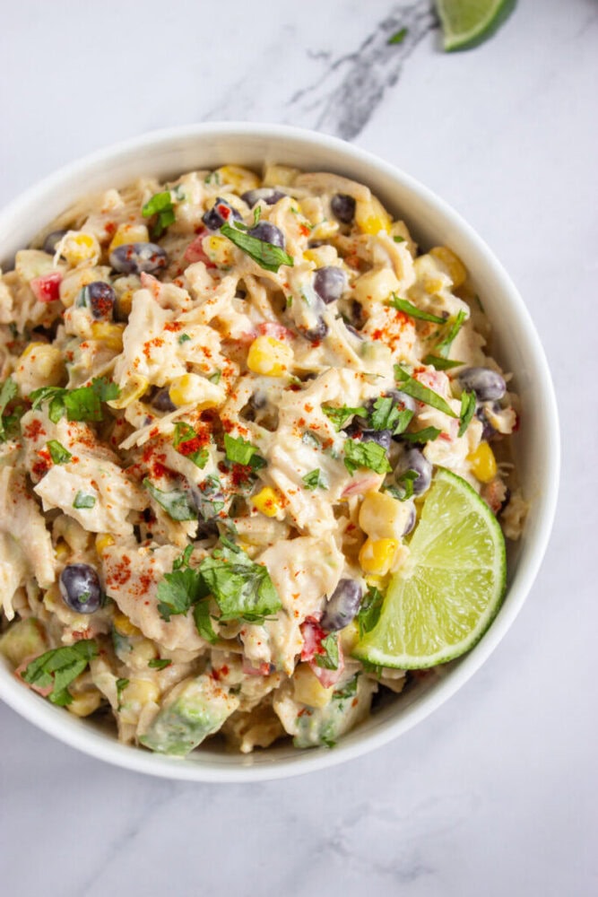 A bowl of creamy chicken salad with corn, black beans, chopped herbs, and a lime wedge garnish, sprinkled with paprika, on a white surface.