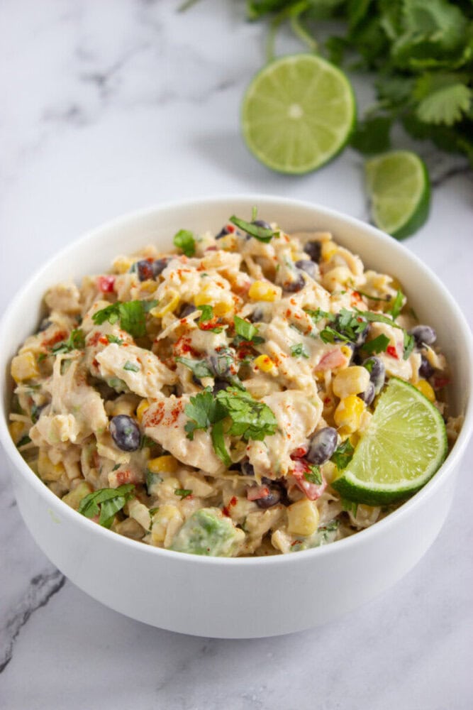 A bowl of creamy salad with black beans, corn, red pepper, chopped cilantro, and a lime wedge, set on a marble surface with lime slices and cilantro in the background.