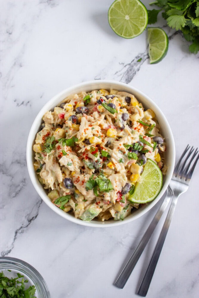 A bowl of creamy pasta salad with black beans, corn, avocado, and herbs, garnished with lime, sits on a marble surface next to forks and fresh cilantro.