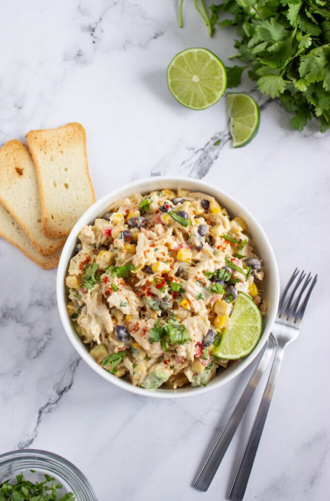A bowl of creamy chicken salad with black beans, corn, and herbs, garnished with a lime wedge, next to toast slices, fresh cilantro, and lime halves on a marble surface.