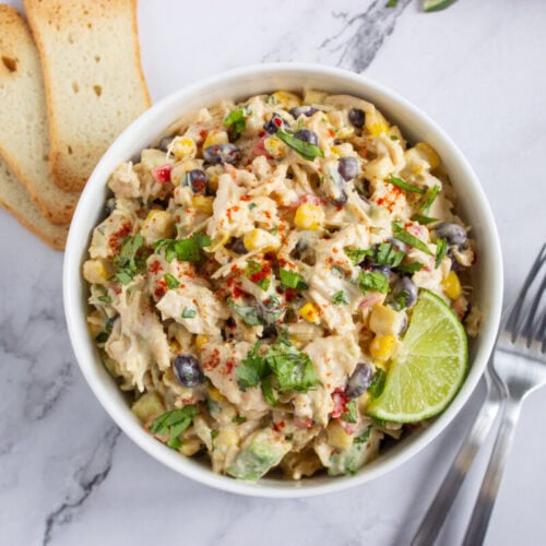 A bowl of creamy chicken salad with corn, black beans, herbs, and lime, served with two toast slices on a marble surface beside two forks.