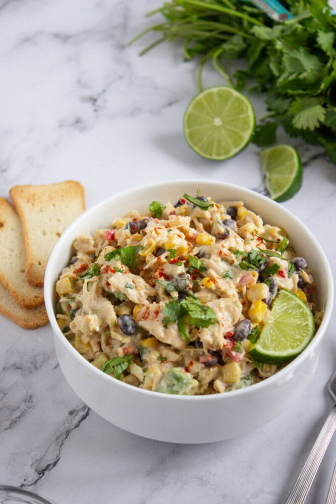 A white bowl of creamy salad with black beans, corn, and chopped herbs, garnished with a lime wedge. Toasted bread slices, lime halves, and fresh cilantro are nearby on a marble surface.