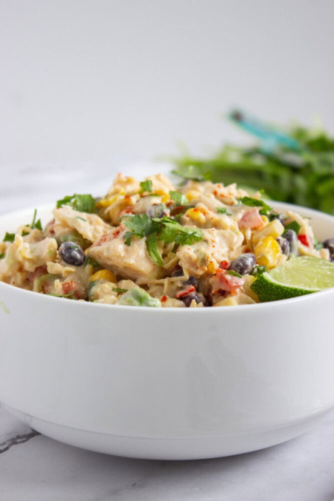 A white bowl filled with a creamy salad containing black beans, corn, chopped vegetables, and garnished with cilantro and a lime wedge.