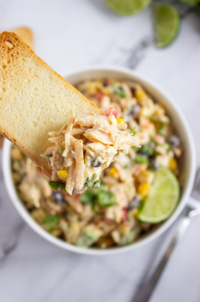 A slice of bread topped with creamy chicken salad, held above a bowl filled with the same salad, garnished with a lime wedge.