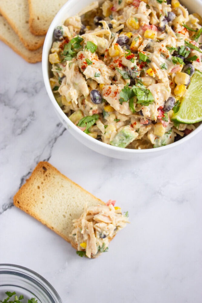 A bowl of chicken salad with black beans, corn, and herbs sits on a marble surface next to slices of bread, one topped with the salad.