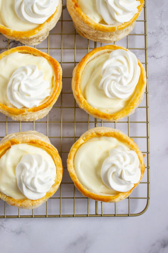 Six small pastry tarts filled with cream and topped with a swirl of whipped cream, arranged on a wire cooling rack.