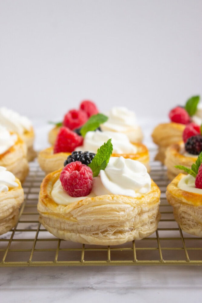 Puff pastry rounds topped with whipped cream, fresh raspberries, blackberries, and mint leaves, arranged on a cooling rack.