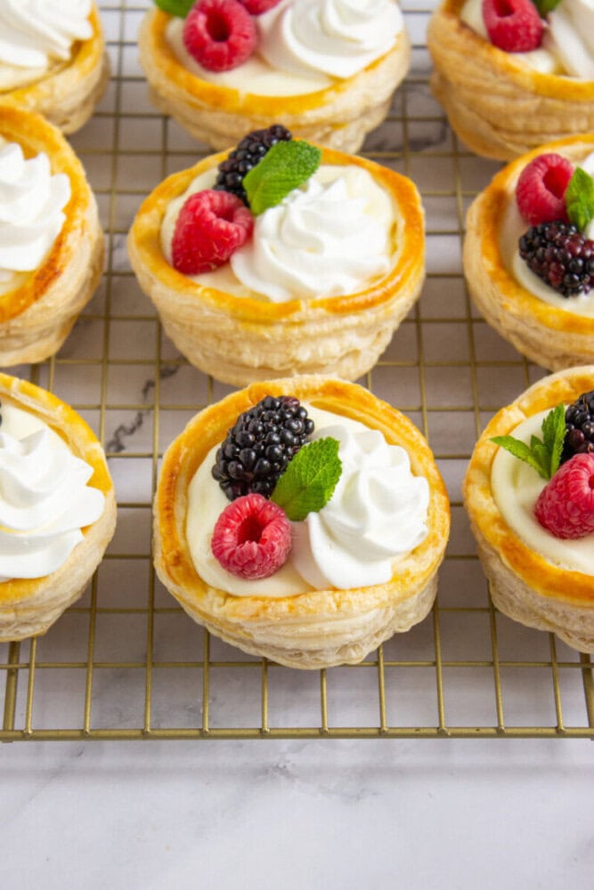 Mini pastry tarts with whipped cream, raspberries, blackberries, and mint leaves are arranged on a cooling rack.