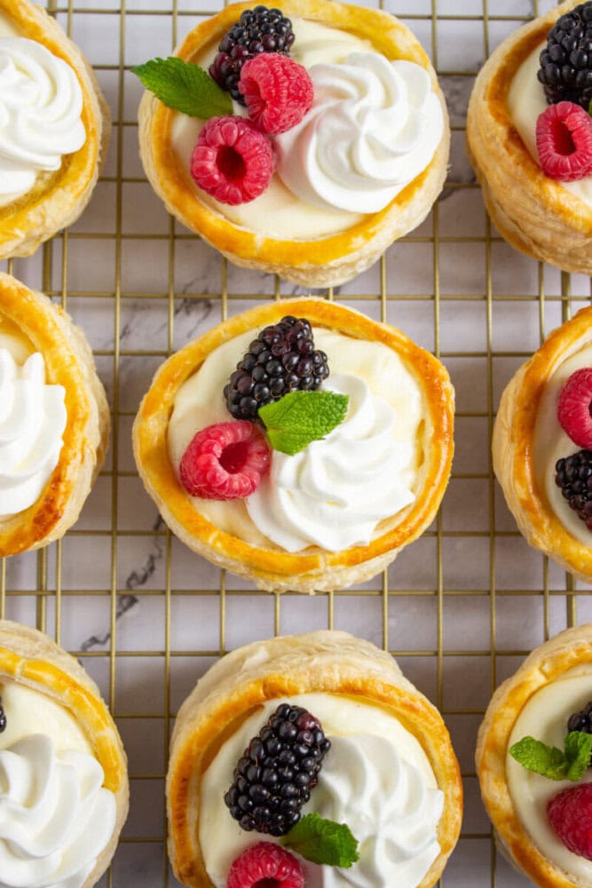 Round puff pastry tartlets filled with cream, topped with whipped cream, fresh raspberries, blackberries, and mint leaves, arranged on a wire cooling rack.