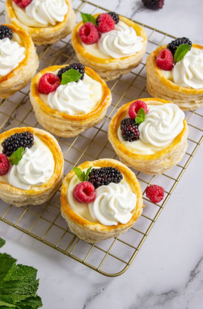 Mini pastry tarts topped with whipped cream, fresh raspberries, blackberries, and mint leaves are arranged on a cooling rack.