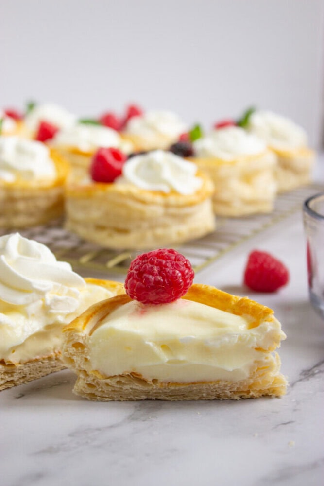 A puff pastry tart filled with cream, topped with whipped cream and a raspberry, is cut in half, with more tarts and raspberries in the background.