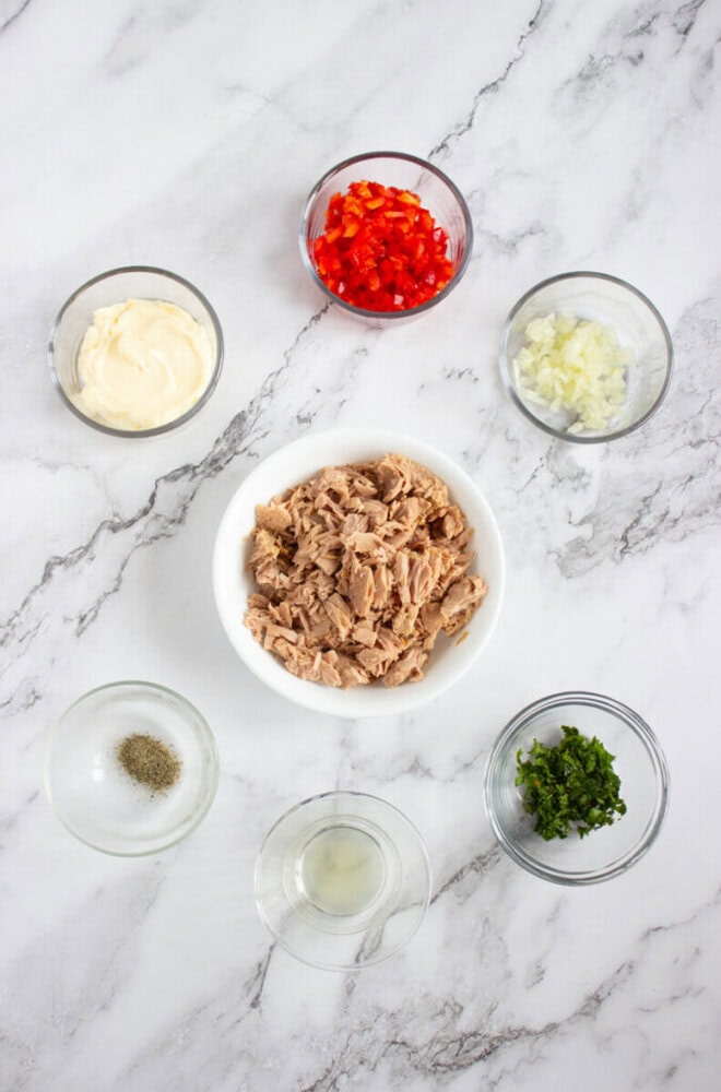 Six small bowls on a marble surface containing tuna, chopped red bell pepper, diced onion, chopped herbs, mayonnaise, ground pepper, and lemon juice.