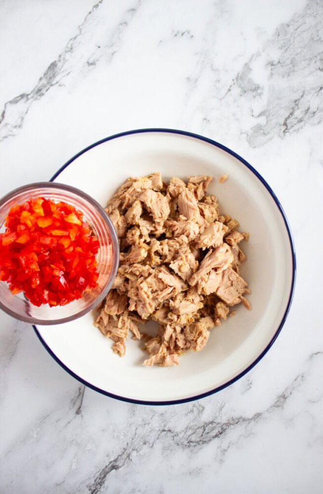 A bowl with chunks of tuna and a smaller bowl containing diced red bell pepper on a marble surface.