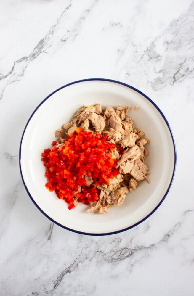 A white bowl containing chunks of tuna and a pile of finely chopped red bell pepper on a marble surface.