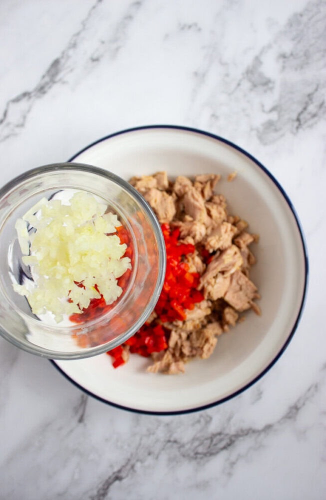 A bowl with chopped tuna and red bell pepper, with a glass container of diced onions being added on a marble countertop.