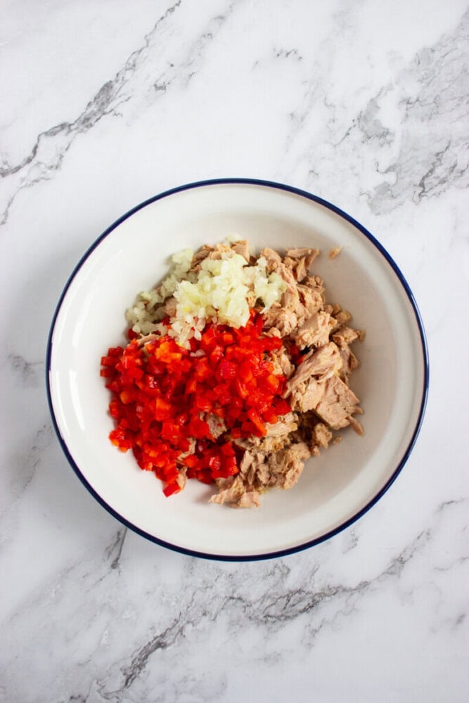 A white bowl containing chopped onions, diced red bell peppers, and flaked tuna on a marble surface.