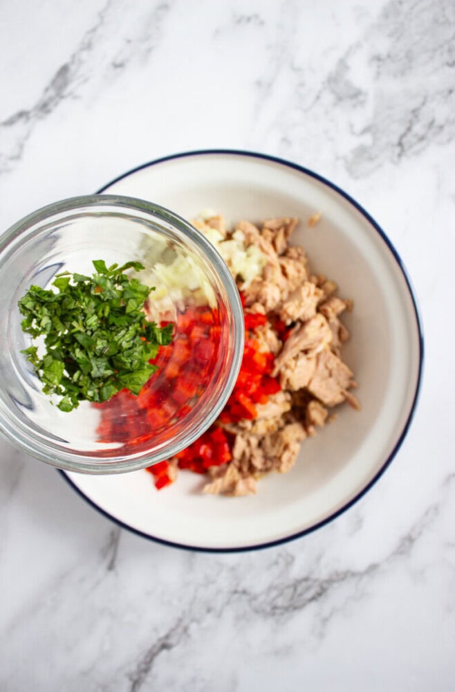 A bowl with tuna, chopped red bell pepper, and onions, with a glass being poured in containing chopped herbs, on a marble surface.