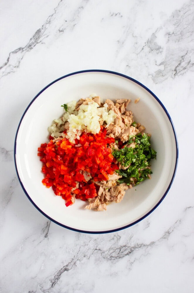 A white bowl on a marble surface containing chopped red bell pepper, onion, green herbs, and chunks of tuna.