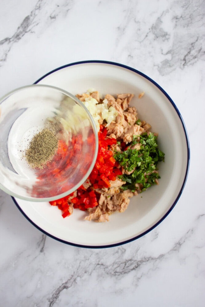 A white bowl with chopped red bell pepper, onion, cilantro, and tuna. A hand pours ground black pepper from a glass container into the bowl.