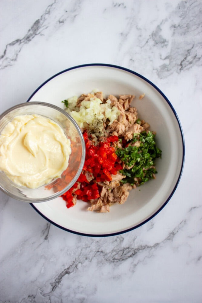 A bowl with chopped onions, red peppers, parsley, and tuna beside a glass of mayonnaise on a marble surface.