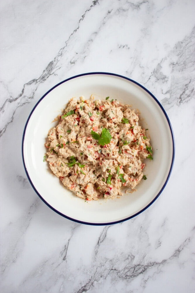 A white bowl with a blue rim filled with a tuna salad mixture, garnished with chopped cilantro, on a white marble surface.