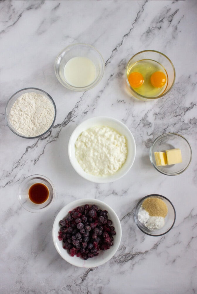 Eight bowls on a marble surface containing flour, vanilla extract, milk, cottage cheese, eggs, butter, blueberries, and a mix of sugar and baking powder.