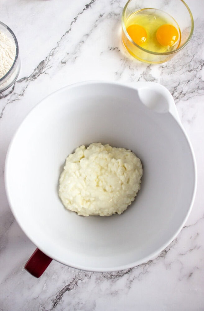 A white mixing bowl with a portion of cottage cheese, next to a bowl containing two cracked eggs and a partial view of a bowl of flour on a marble countertop.