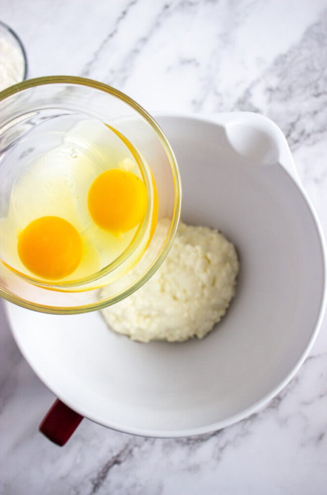 A glass bowl with two raw eggs is held above a white mixing bowl containing a mound of ricotta cheese, on a marble countertop.